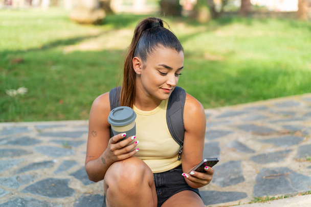 Joven mujer bonita al aire libre usando el teléfono móvil y sosteniendo un café para llevar - Foto, Imagen
