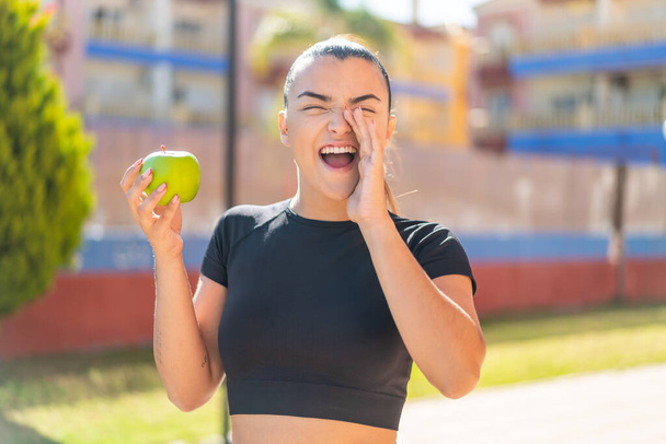 Joven mujer morena bonita con una manzana al aire libre gritando con la boca abierta - Foto, Imagen