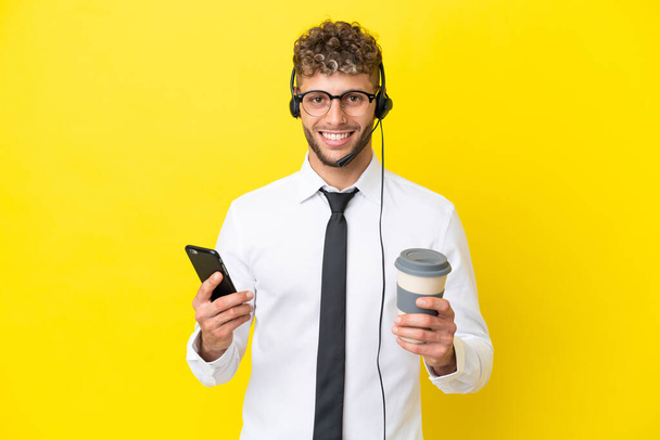 Hombre rubio telemarketer trabajando con un auricular aislado sobre fondo amarillo sosteniendo café para llevar y un móvil - Foto, imagen