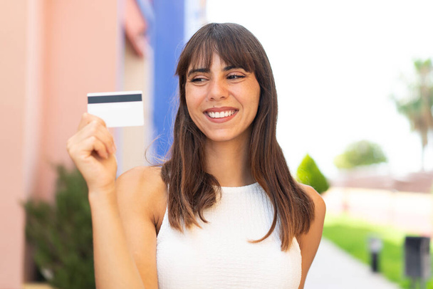 Mladá žena drží kreditní kartu venku s veselým výrazem - Fotografie, Obrázek