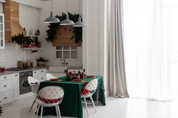 кухня, прикрашена сосновими гірляндами та різдвяними іграшками. Стіл і поруч стоїть ялинка. інтер'єр яскравої кухні прикрашений на Різдво
 - Фото, зображення