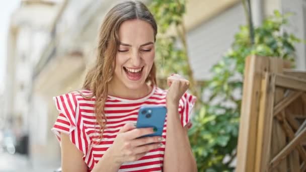 Mujer rubia joven usando smartphone con gesto ganador en la terraza de la cafetería - Imágenes, Vídeo