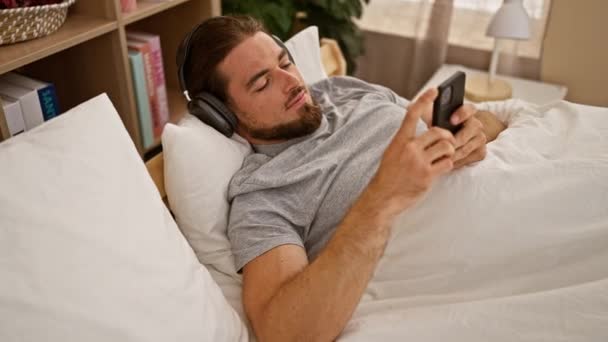 Joven hombre hispano usando smartphone acostado en la cama usando auriculares en el dormitorio - Imágenes, Vídeo