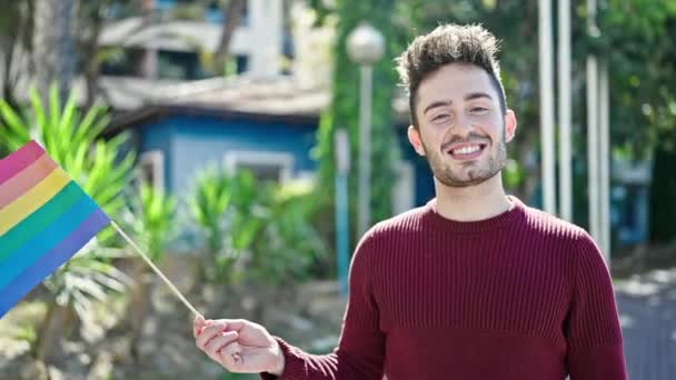 Joven hombre hispano sonriendo confiado sosteniendo bandera de arco iris en el parque - Imágenes, Vídeo