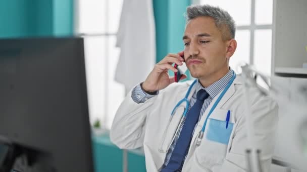 若いヒスパニック系の男の医者を使用してコンピュータを使用してスマートフォンで話すクリニック - 映像、動画