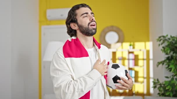 Joven hispano cantando himno del equipo de fútbol con la mano en el corazón sosteniendo la pelota de fútbol en casa - Metraje, vídeo