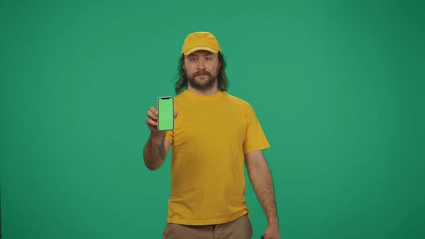 Portrait of a delivery man in yellow cap and tshirt holding a smartphone. Isolated on green background. Advertising area, workspace mockup. - Foto, imagen