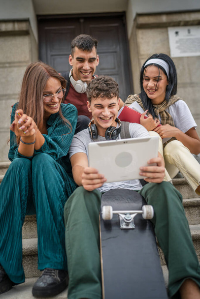 Group of caucasian teenage friends sit in front of school or university hold digital tablet watch video movie or have video call browse internet online for social network real people - Photo, Image
