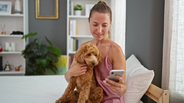 Giovane donna caucasica con cane utilizzando smartphone seduto sul letto in camera da letto - Filmati, video