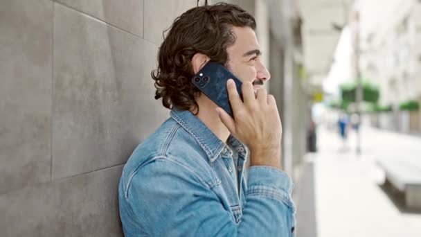 Joven hombre hispano sonriendo confiado hablando en el teléfono inteligente en la calle - Metraje, vídeo