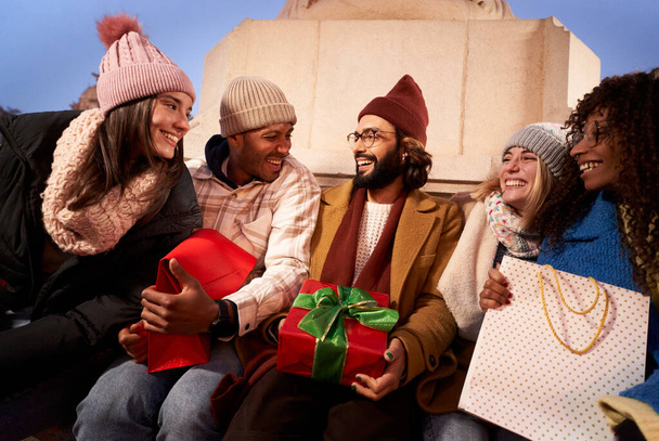 Glimlachende multiraciale groep mensen zitten met de huidige winter kerstcadeaus. Vrolijke jonge vrienden genieten samen van een vreugdevolle buitenavond. Communautaire betrekkingen, vrijetijdsbesteding en verjaardagen. - Foto, afbeelding