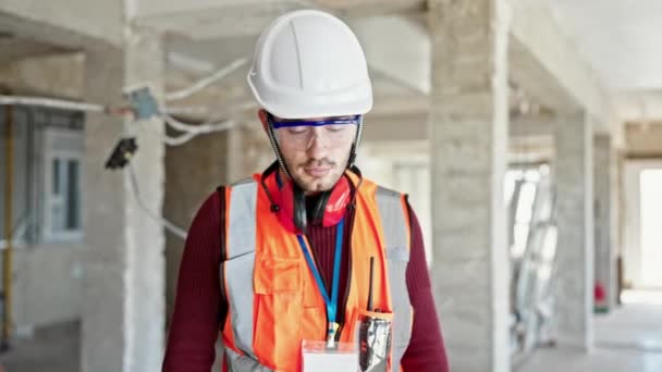 Joven constructor hispano sonriendo confiado de pie en el sitio de construcción - Imágenes, Vídeo