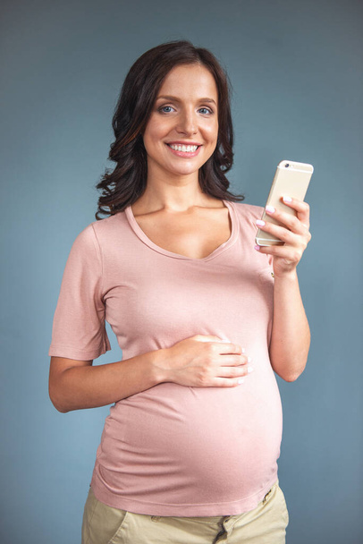カジュアルな服で美しい妊娠中の若い女性がスマート フォンを使用して、灰色の背景に、笑みを浮かべて彼女のおなかに触れて - 写真・画像