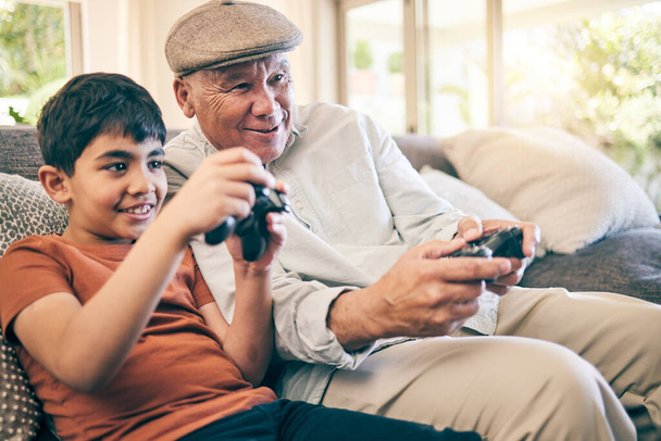 Οικογένεια, διασκέδαση και ένα αγόρι που παίζει με τον παππού του σε έναν καναπέ στο σαλόνι του σπιτιού τους κατά τη διάρκεια μιας επίσκεψης. Βιντεοπαιχνίδι, παιδιά κι ένας ηλικιωμένος μαθαίνουν να παίζουν από τον εγγονό του.. - Φωτογραφία, εικόνα