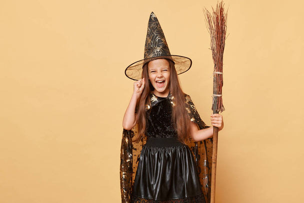 Spooky witchcraft tradition. Magical wizard and sorcerer. Crazy excited little girl with long hair black cloak clothing with broom isolated over beige background - Photo, image
