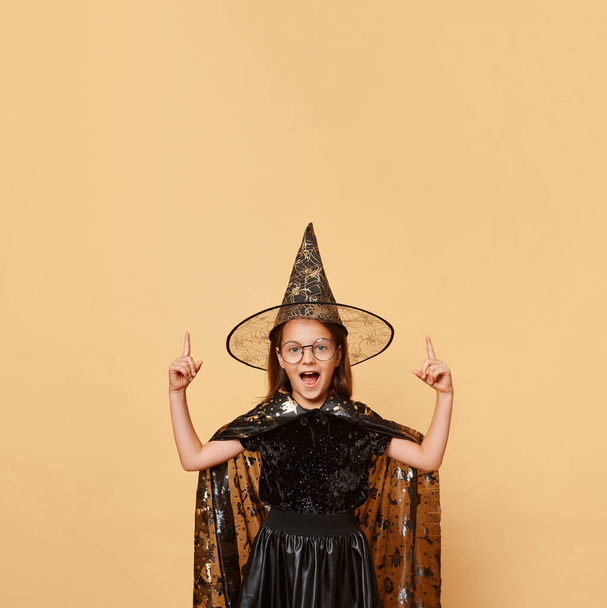 Excited crazy little girl wearing witch costume carnival cone hat and glasses isolated over beige background screaming with amazement pointing up at advertisement area. - Foto, immagini