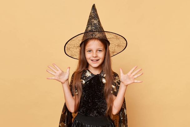Cute little girl sorcerer wearing witch costume and carnival cone hat isolated over beige background raised her arms making halloween magic spell with happy face - Fotoğraf, Görsel