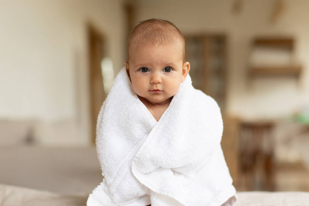 Yatma vakti. Sevimli küçük bebek beyaz havluya sarılmış bir şekilde yatakta oturuyor, kameraya bakıyor, boşluğu kopyalıyor. - Fotoğraf, Görsel