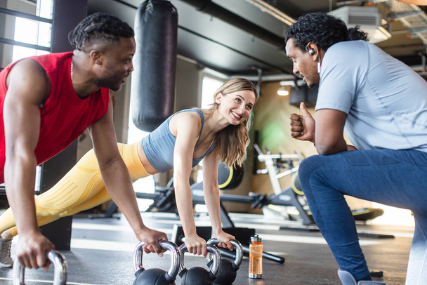 Diversity Frau und Mann in modernem Fitnessstudio machen Liegestütze mit Wasserkocher unter Aufsicht des Trainers - Foto, Bild