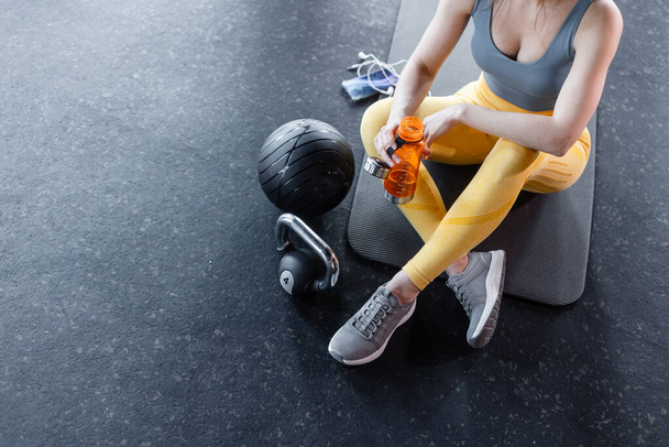 fit a krásná mladá žena ve sportovním oblečení sedí na podlaze tělocvičny vedle varné konvice zvonek s telefonem a láhev vody v ruce - Fotografie, Obrázek