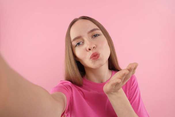Young woman taking selfie and blowing kiss on pink background - Фото, зображення