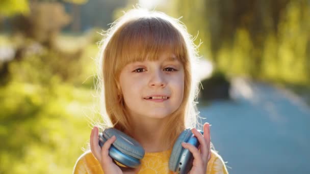 Šťastné usměvavé předpubertální dítě nosí bezdrátové sluchátka výběru, poslouchá energickou disko hudbu v smartphonu tance. Blondýny legrační dívka dívá na kameru stojící na město západ slunce park ulici - Záběry, video