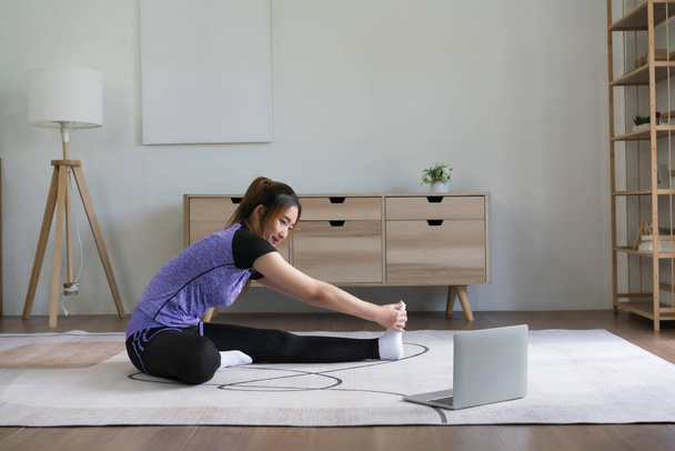 jong aziatische vrouw kijken fitness video op laptop naar training en het doen van oefening naar stretching been. - Foto, afbeelding