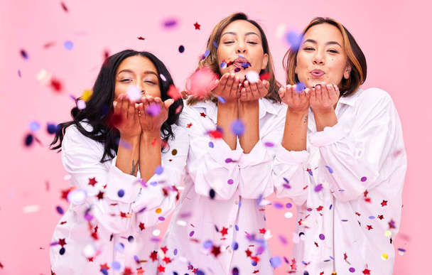 Blowing confetti, friends and women in studio for party celebration, fashion sale and discount. Bonding, diversity and happy people on pink background for support, natural beauty and retail promotion. - Photo, Image