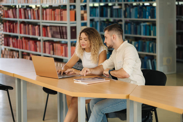 Üniversite kütüphanesinde okuyan iki kız ve erkek arkadaş. Laptop kullanan bir kız. Kitapların arkaplanı. Yüksek kalite fotoğraf - Fotoğraf, Görsel