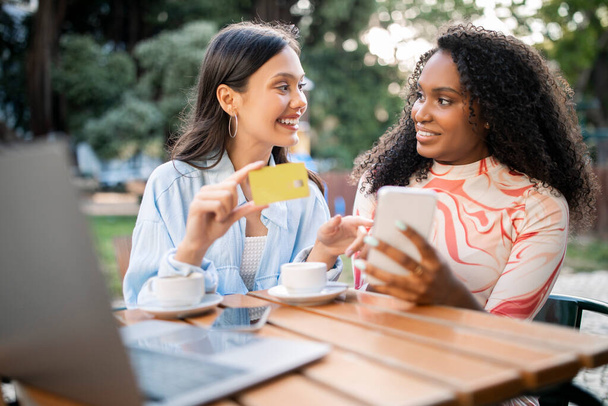 Две счастливые подруги с помощью смартфона и кредитной карты, сидя в кафе на открытом воздухе, молодые веселые многоэтнические Besties делает онлайн-платежей или покупки в Интернете, наслаждаясь электронной коммерции - Фото, изображение