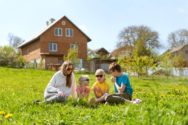 Anne ya da dadı çayırda çocuklarla oturuyor. Etrafı karahindiba dolu. İki katlı bir eve karşı çimenlerde oturan genç bir aile. - Fotoğraf, Görsel
