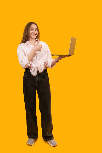 Jeune femme caucasienne tient l'ordinateur portable dans la main et montre pouces vers le haut. Studio intérieur tourné isolé sur fond jaune. Cadre vertical. Espace de copie - Photo, image