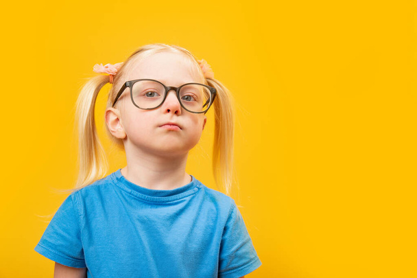 Πορτρέτο του μικρού ξανθού κοριτσιού με δύο ουρές φοράει γυαλιά και κοιτάζει επίμονα στο βάθος. Προσωπογραφία του προσχολικού σε κίτρινο φόντο - Φωτογραφία, εικόνα