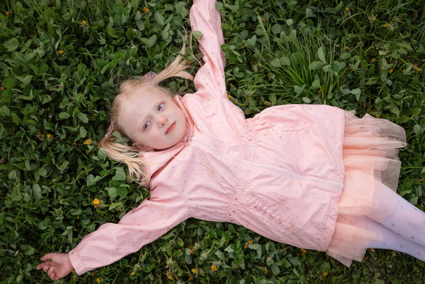 Niña rubia en vestido rosa y chaqueta está acostada sobre hierba verde con los brazos extendidos a los lados. Primavera. Vista superior - Foto, Imagen