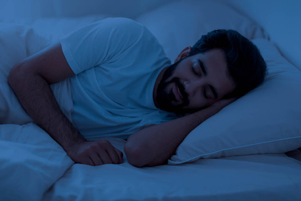Healthy Sleeping Concept. Portrait of young indian man lying in bed with closed eyes in the night, calm eastern male resting in bedroom, relaxing in the dark room, having hap with hand under head - Photo, Image