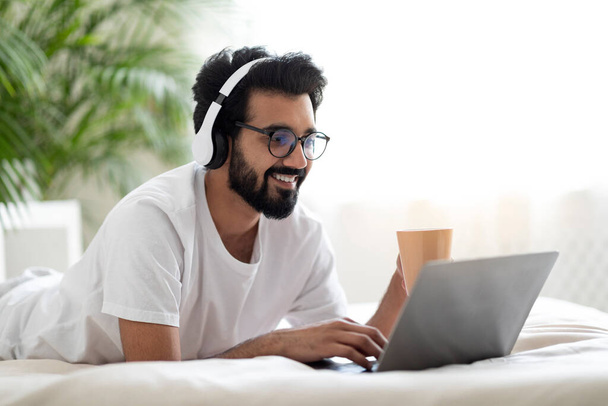Młody Indianin noszący słuchawki bezprzewodowe odpoczywający w łóżku z laptopem i kawą, szczęśliwy uśmiechnięty wschodni człowiek surfujący po Internecie na komputerze i cieszący się gorącym napojem, widok z boku z kopią przestrzeni - Zdjęcie, obraz