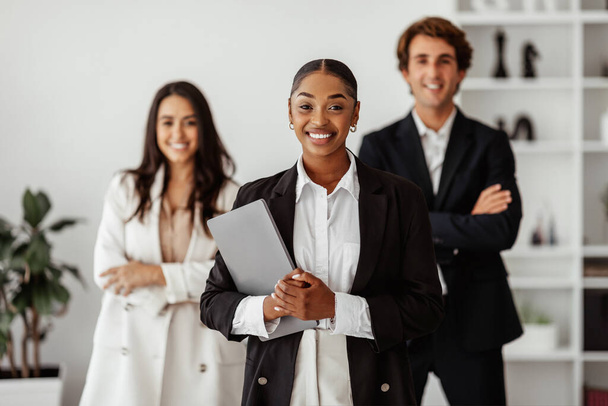 Successful black businesswoman holding laptop in hands, standing with multiracial business collegues in office. Entrepreneurship, career growth, motivation concept - Photo, Image