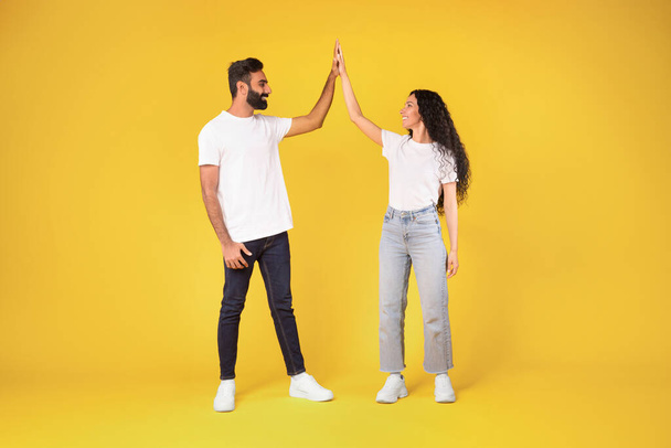 Full Length Of Happy Young Arabic Couple Giving High Five To Each Other Standing Over Yellow Studio Background, Cheerful Middle Eastern Spouses Enjoying Teamwork, Celebrating Common Success - Photo, Image