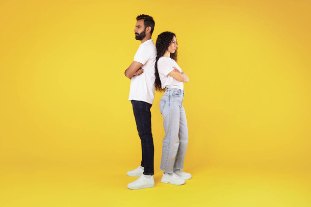 Relationship crisis, disagreement. Offended arab young spouses standing back to back over yellow background in studio, ignoring each other after quarrel, suffering marriage problems. Full length - Photo, Image
