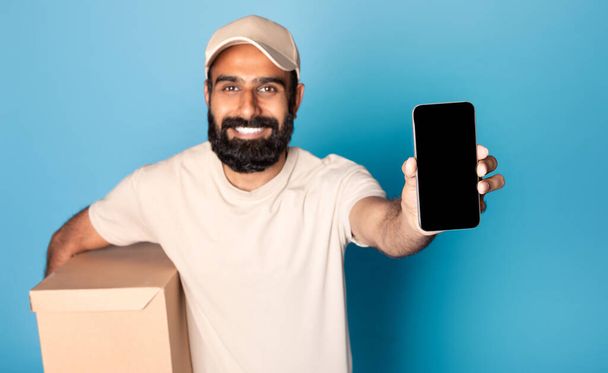 Aplicación de servicio de entrega. Hombre indio mostrando teléfono inteligente con pantalla en blanco para maqueta, llevando uniforme y sosteniendo la caja de cartón, aislado sobre fondo azul - Foto, Imagen