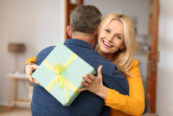 Érzelmes, érett nő ölelgeti a férjét ajándékdobozzal a kezében, megünnepli a házassági évfordulóját a házban, a kamerába mosolyog. A férfi gratulál a nőnek, meglepi őt ajándékkal a nyaraláson - Fotó, kép