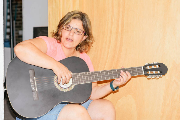 femme jouant sa chanson préférée à la guitare acoustique noire, assise sur le canapé à la maison. Loisirs musicaux, temps libre. - Photo, image