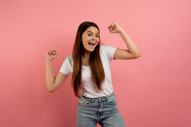 Hooray. Overjoyed teen girl shaking clenched fists, charing and rejoicing success, happy excited female teenager celebrating win while standing over pink studio background, copy space - Photo, Image
