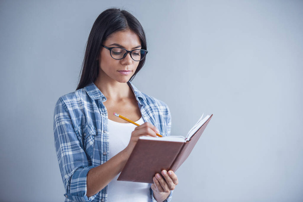 schönes junges Mädchen in Freizeitkleidung und Brille schreibt in ihr Notizbuch, auf grauem Hintergrund - Foto, Bild
