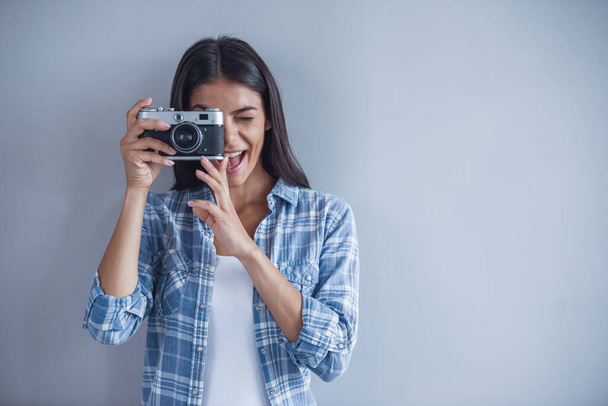 Beautiful young girl in casual wear is making photos using a camera and smiling, on gray background - Photo, Image