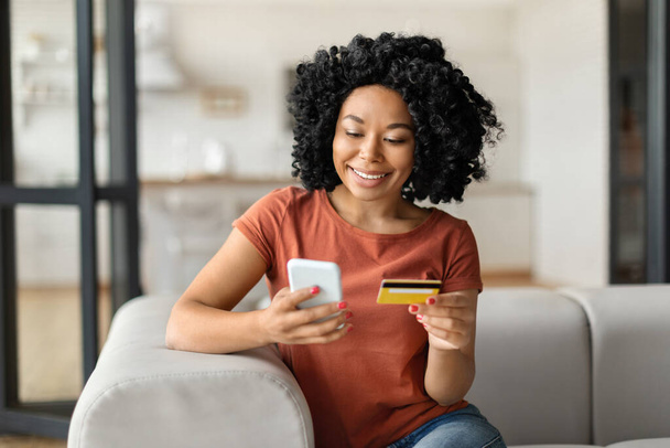 Happy Young Black Woman Zakupy online ze smartfonem i kartą kredytową podczas relaksu na kanapie w domu, Uśmiechnięta Afroamerykanka za pomocą e-commerce na zakupy internetowe, Kopiuj przestrzeń - Zdjęcie, obraz