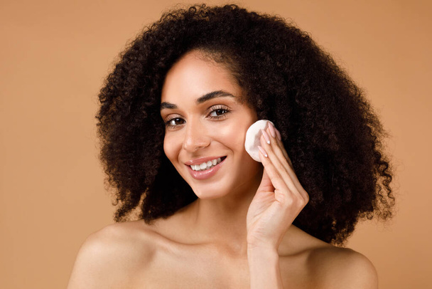 Daily skincare routine. Young lady cleansing skin with cotton pad, latin woman removing makeup from face, standing over brown studio background and smiling at camera - Photo, Image