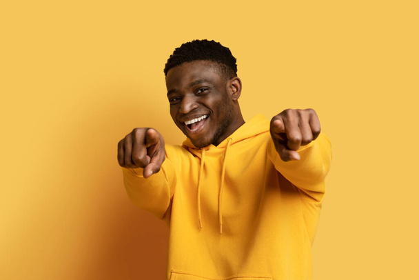 Pozytywny, pogodny, przystojny, milenijny, afrykański facet w żółtej bluzie z kapturem, gestykulujący obiema rękami i uśmiechający się do kamery, odizolowany na żółtym tle studia - Zdjęcie, obraz