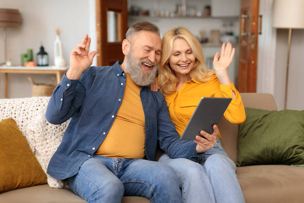 Vanhukset virtuaalisessa viestinnässä. Onnellinen kypsä pari tekee videopuhelun digitaalisen tabletin tietokoneen avulla, tervehtimällä ja kommunikoimalla verkossa istuen yhdessä sohvalla kotona - Valokuva, kuva