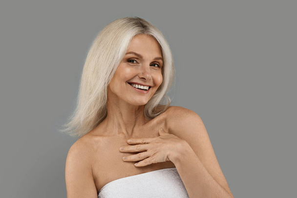 Aged Beauty. Portrait Of Attractive Smiling Mature Woman Wrapped in Towel Posing Over Grey Studio Background, Happy Beautiful Elderly Lady Touching Her Chest And Looking At Camera, Copy Space - Photo, Image